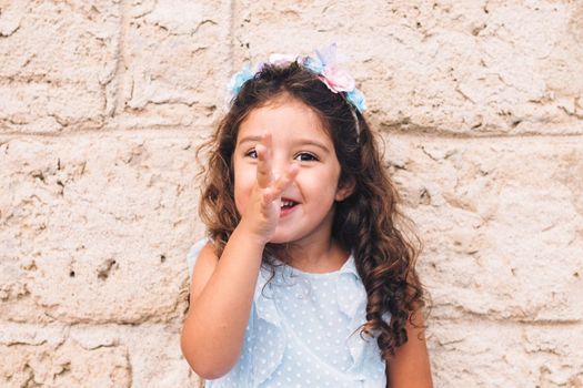 little girl making fun with her hand on her nose, is in front of a stone wall and wears a blue dress and a flower headband