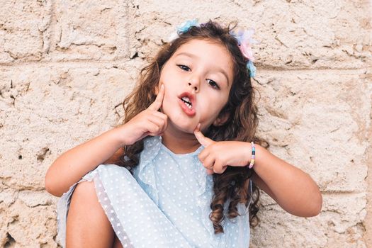 little girl making fun with hands and face, is in front of a stone wall and wears a blue dress and a flower headband