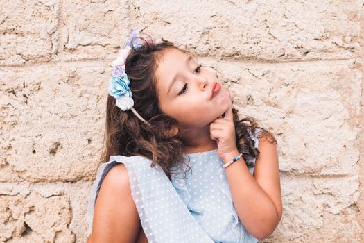 funny little girl doing like she thinks, is in front of a stone wall and wears a blue dress and a flower headband