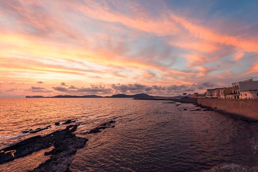 Spectacular and colorful sunset in Alghero, Sardinia, Italy. On one side is the seafront of the city and in the background the cape of the giant that sleeps