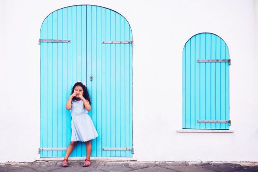 funny little girl making fun with her eyes, she is in front of a white wall with a door and a turquoise wooden window