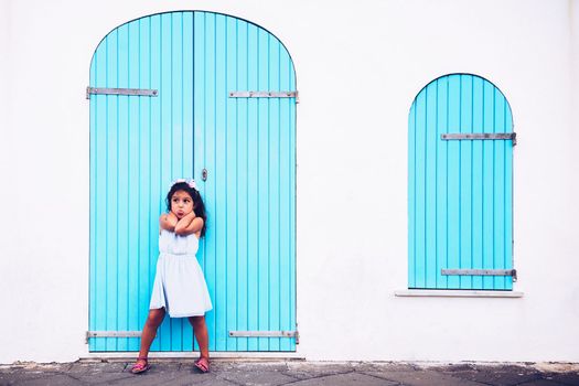 funny little girl making fun by sticking out her tongue, she is in front of a white wall with a door and a turquoise wooden window
