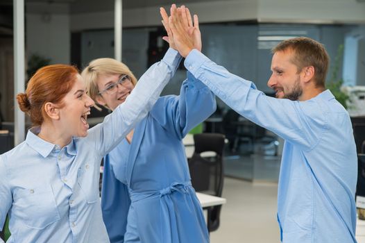 Colleagues give a high five in the office. Red-haired woman, blonde and bearded man in a denim shirt in the office