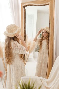 A beautiful teenage girl with long hair measures a straw hat in front of a mirror. Self-admiration of a blonde. selective focus