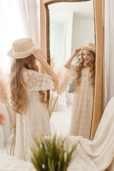 A beautiful teenage girl with long hair measures a straw hat in front of a mirror. Self-admiration of a blonde. selective focus