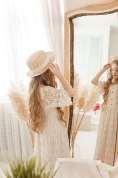 A beautiful teenage girl with long hair measures a straw hat in front of a mirror. Self-admiration of a blonde. selective focus