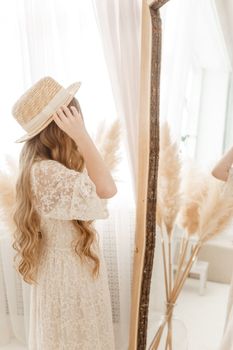 A beautiful teenage girl with long hair measures a straw hat in front of a mirror. Self-admiration of a blonde. selective focus