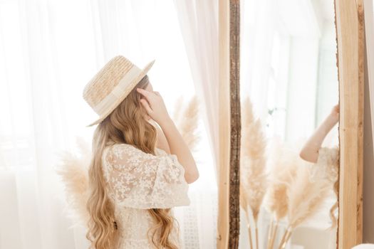 A beautiful teenage girl with long hair measures a straw hat in front of a mirror. Self-admiration of a blonde. selective focus