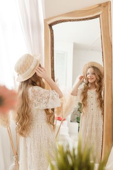 A beautiful teenage girl with long hair measures a straw hat in front of a mirror. Self-admiration of a blonde. selective focus