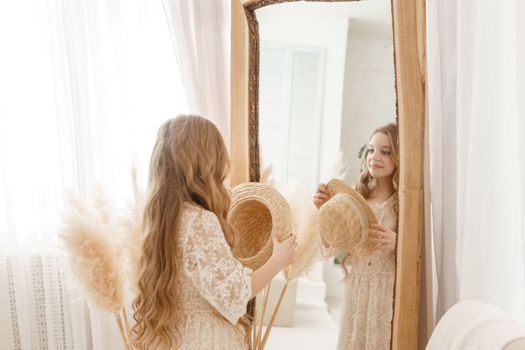 A beautiful teenage girl with long hair measures a straw hat in front of a mirror. Self-admiration of a blonde. selective focus