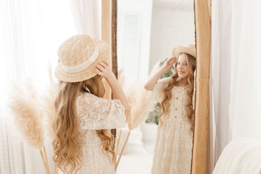 A beautiful teenage girl with long hair measures a straw hat in front of a mirror. Self-admiration of a blonde. selective focus