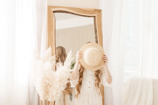 A beautiful teenage girl with long hair measures a straw hat in front of a mirror. Self-admiration of a blonde. selective focus