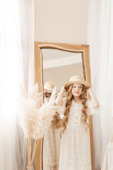 A beautiful teenage girl with long hair measures a straw hat in front of a mirror. Self-admiration of a blonde. selective focus