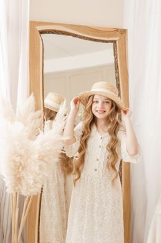 A beautiful teenage girl with long hair measures a straw hat in front of a mirror. Self-admiration of a blonde. selective focus