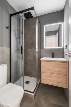 Small bathroom in a modern design with bright lighting. Shower cabin made of ergonomic glass with black faucet. Vanity table with wooden drawers and white sink is in harmony with overall design