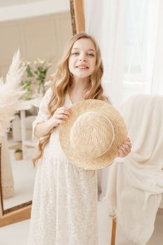 A beautiful teenage girl with long hair measures a straw hat in front of a mirror. Self-admiration of a blonde. selective focus