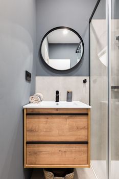 Bathroom with a washbasin on a floating vanity. On a gray wall is a round mirror in a black metal frame. Bathroom is filled with a fashionable design solution, pleasant and comfortable little things