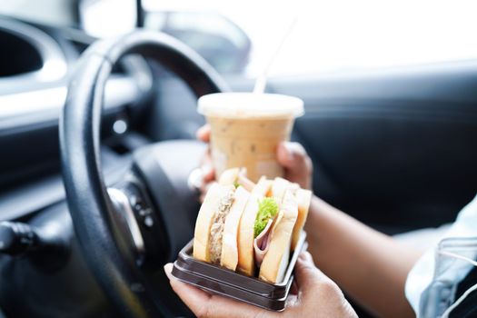 Asian woman driver hold ice coffee cup and sandwich bread for eat and drink in car, dangerous and risk an accident.