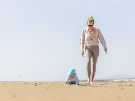 Mother playing his infant baby boy son on sandy beach enjoying summer vacationson on Lanzarote island, Spain. Family travel and vacations concept.