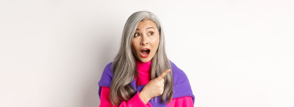 Close-up of amazed and intrigued asian senior woman pointing finger left at something cool, staring at camera promo offer, standing over white background.