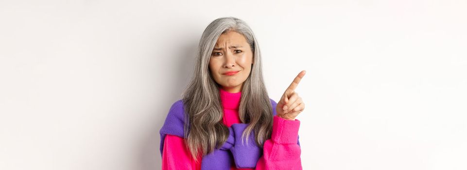 Judgy and skeptical asian senior woman grimacing displeased, pointing finger upper left corner unamused, standing over white background.