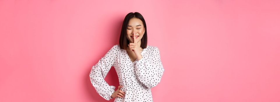 Beautiful Korean woman in trendy dress asking to keep secret, hushing with soft smile and closed eyes, standing over pink background.