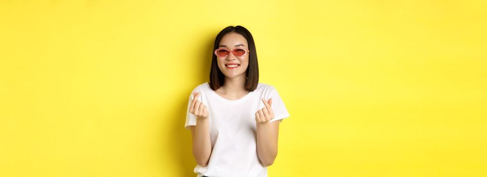 Fashion and lifestyle concept. Attractive asian woman in stylish sunglasses, showing finger hearts and smiling happy at camera, standing over yellow background.