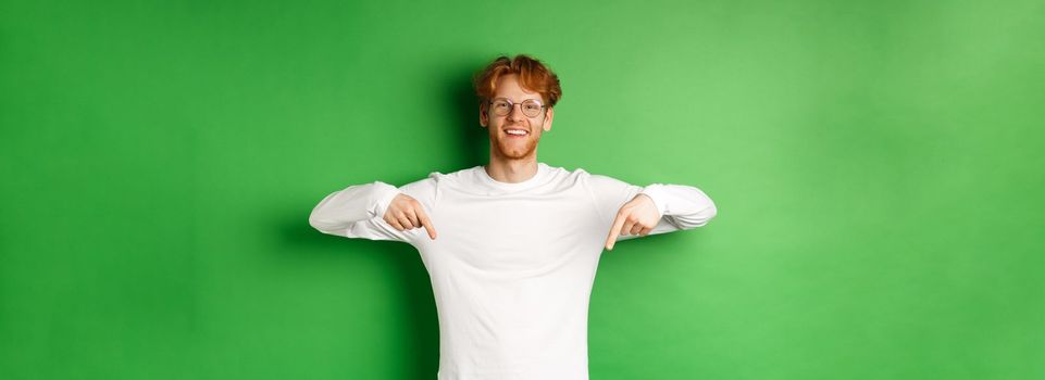 Handsome male model with red hair, wearing glasses and long-sleeve, pointing fingers down and smiling cheerful, showing promo, green background.
