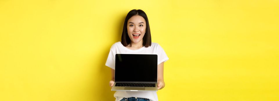 Young asian woman demonstrate online offer, showing blank laptop screen and smiling, standing over yellow background.