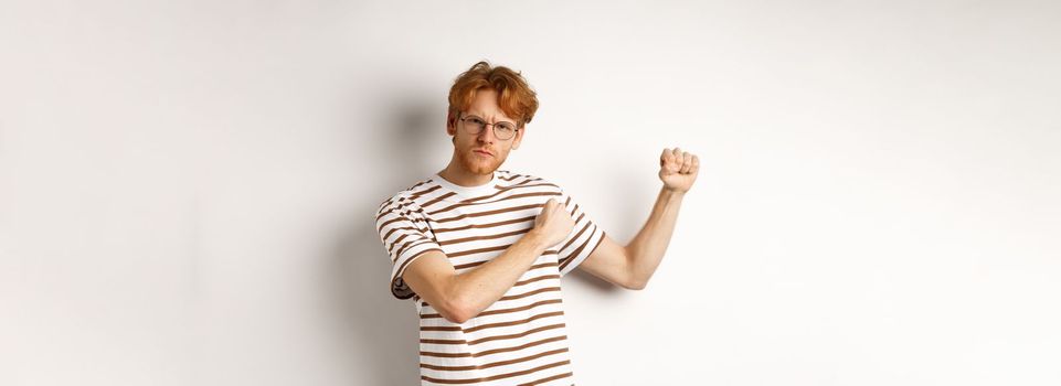 Serious and confident redhead man in glasses raising fists, ready for fight, staring angry at camera while shadow boxing, standing over white background.