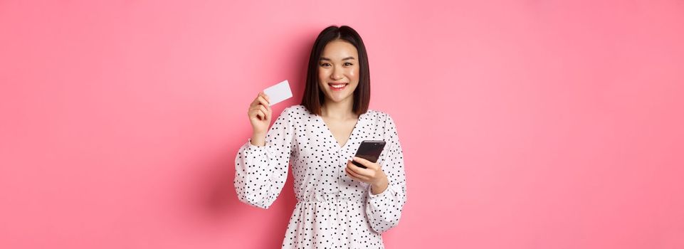 Attractive young asian woman order online, holding credit card and mobile phone, making internet purchase, standing happy over pink background.
