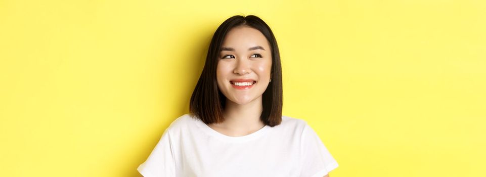 Close up of good-looking asian woman in white t-shirt, smiling and glancing left with pleased face, standing over yellow background.