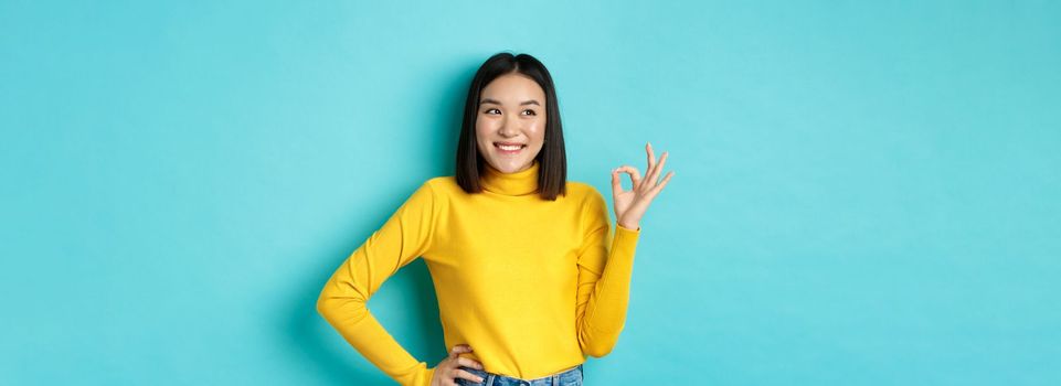 Impressed asian woman in yellow sweater, looking left amazed and showing OK sign in approval, standing over blue background.