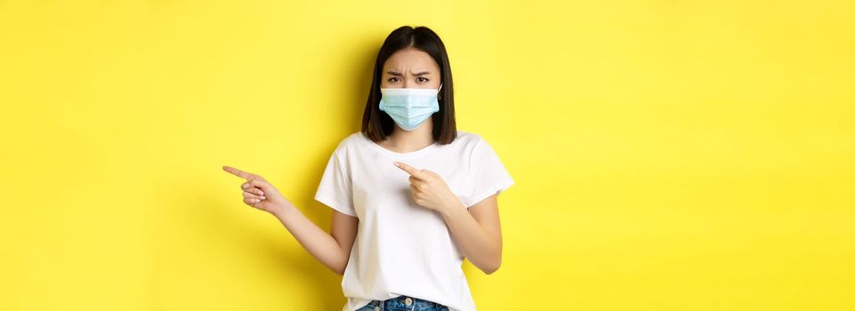 Covid-19, pandemic and social distancing concept. Disappointed asian girl in medical mask, frowning upset and pointing fingers right at logo, standing over yellow background.