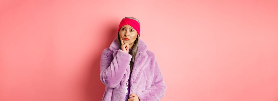 Funky old asian woman looking thoughtful and perplexed, standing in fake fur coat and thinking, making decision, standing on pink background.