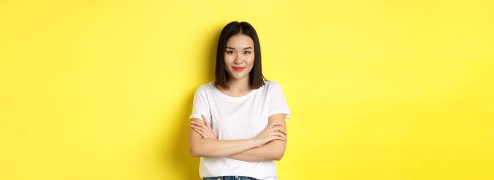 Confident and stylish asian woman cross arms on chest and smiling, standing over yellow background.