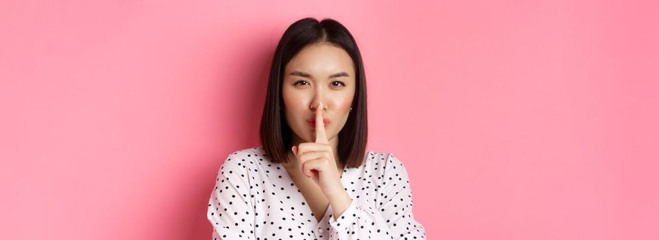 Close-up of mysterious asian woman hiding a secret, hushing and telling to keep quiet, standing over pink background. Copy space