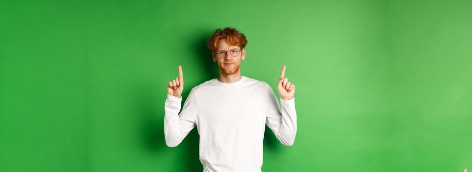 Displeased young man with red hair and glasses, pointing fingers up and frowning doubtful, feeling disappointed, standing over green background.