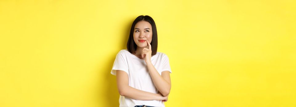 Beauty and fashion concept. Pensive asian woman thinking, looking thoughtful while pondering something, standing over yellow background.