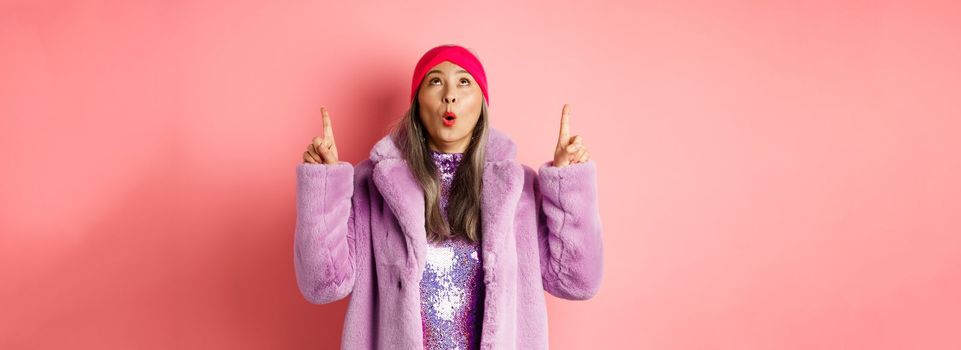 Impressed asian elderly woman in stylish fake-fur coat checking out promo, looking and pointing fingers up, standing over pink background.