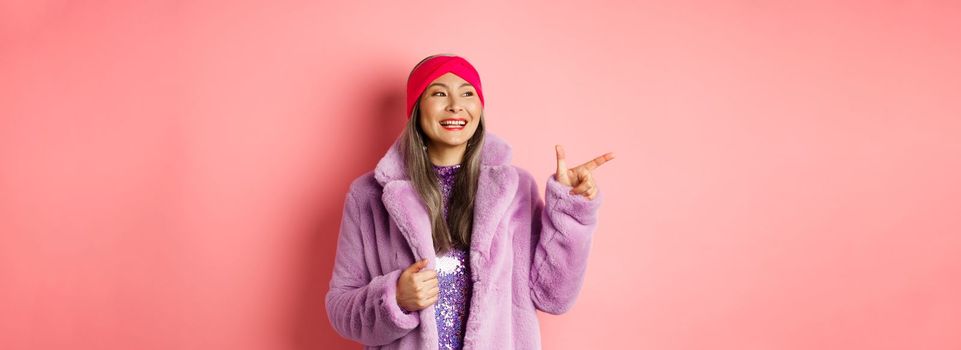 Happy senior asian woman smiling, pointing finger left and looking satisfied, checking out special promotion, pink background.