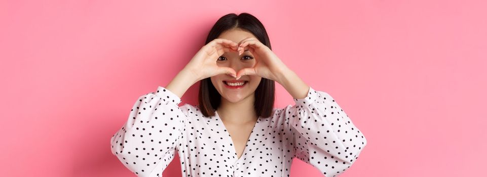 Beauty and lifestyle concept. Close-up of lovely asian woman showing heart sign, smiling and feeling romantic on valentines day, standing over pink background.