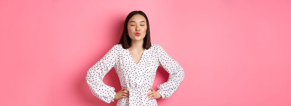 Beautiful asian woman in dress pucker lips and close eyes, waiting for kiss, standing over pink background.