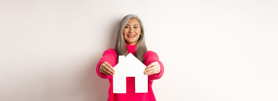 Real estate. Happy korean middle-aged woman with grey hair, showing paper house model and smiling, standing in pink sweater over white background.