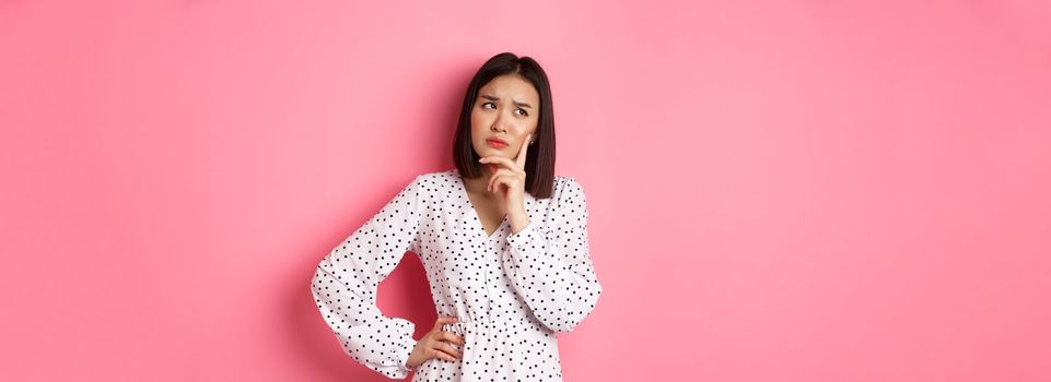 Concerned young asian female model making decision, seriously thinking, looking at upper left corner and choosing, standing over pink background.