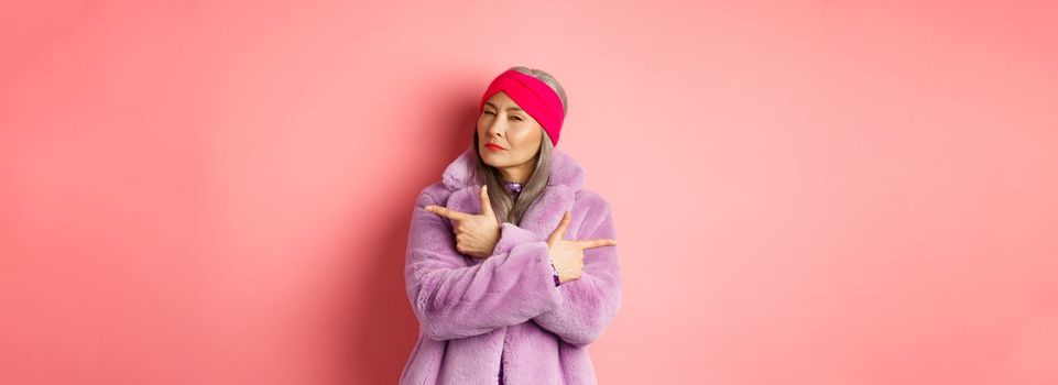Fashion and shopping concept. Stylish asian mature woman making decision, squinting thoughtful while pointing fingers sideways, showing two variants on pink background.