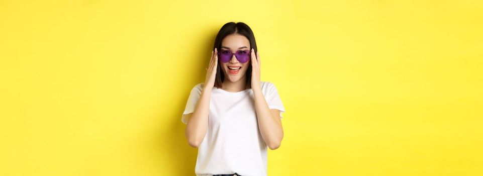 Fashion and lifestyle concept. Stylish asian woman trying new sunglasses in heart-shape, going on vacation, smiling happy at camera, standing over yellow background.