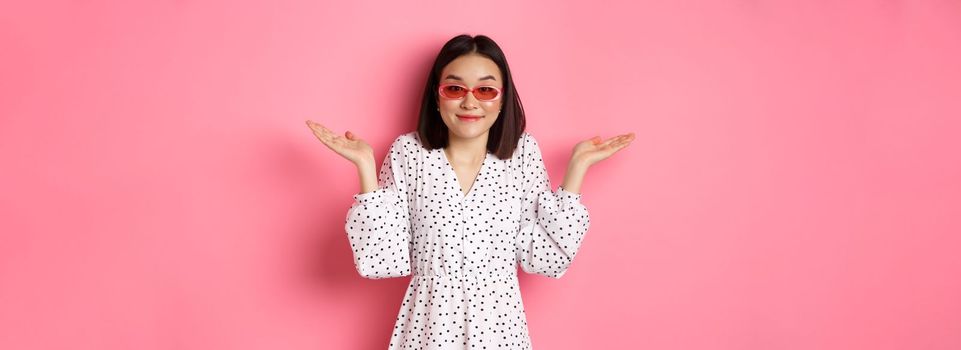 Cute asian woman tourist smiling at camera, shrugging clueless, dont know, wearing trendy sunglasses and white dress, standing against pink background.