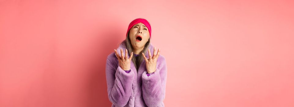Miserable asian senior woman in stylish purple faux fur coat, pleading god and asking why, shaking hands and looking up desperate, standing over pink background.