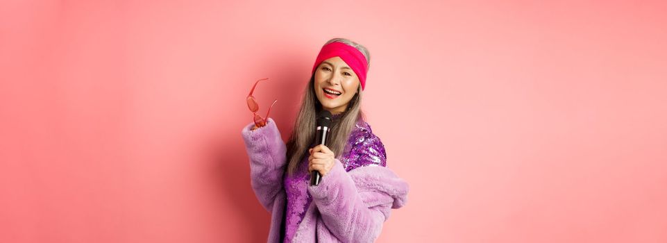Gorgeous asian senior woman singing karaoke in mic, perform song and looking happy, standing over pink background.